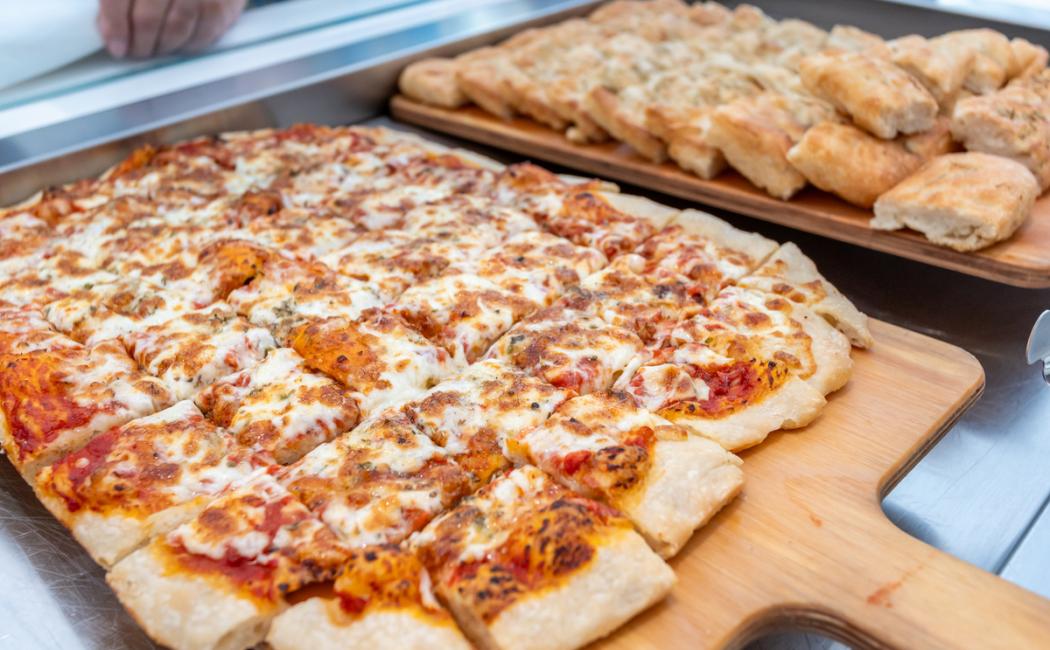 Pizza rettangolare al formaggio e pane focaccia su taglieri di legno.