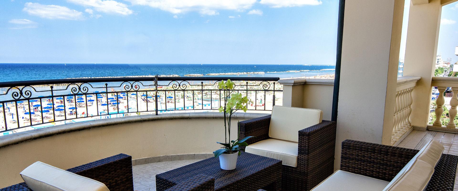 Terrazza con vista mare, arredi in rattan e fiori, spiaggia con ombrelloni.
