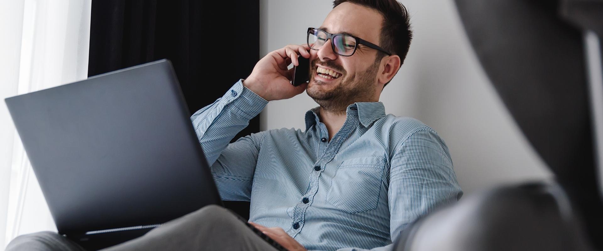 Uomo sorridente al telefono con laptop, indossa camicia a righe e occhiali.