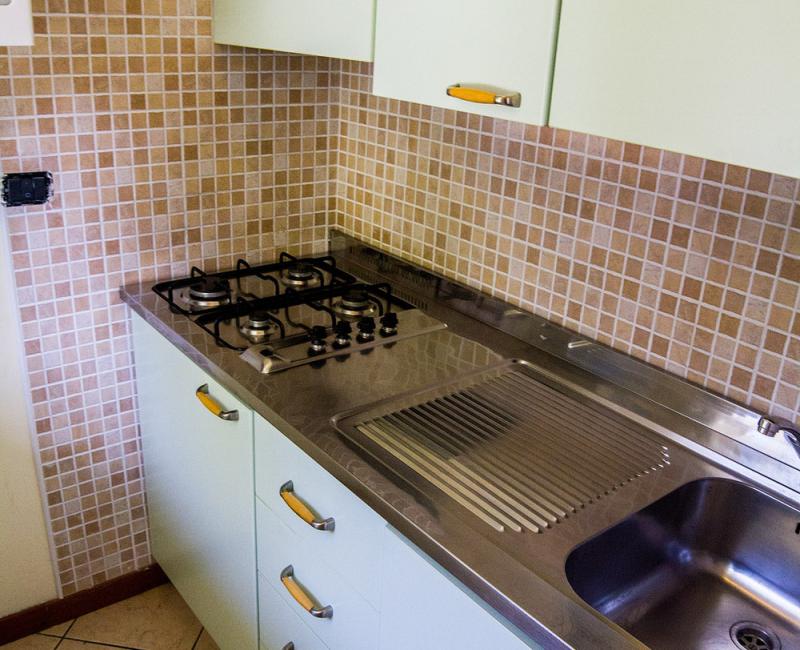 Piccola cucina con fornelli a gas, lavello in acciaio e piastrelle beige.