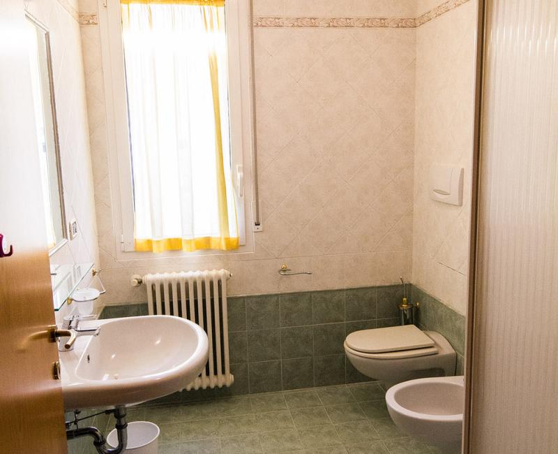 Bagno luminoso con lavabo, bidet, doccia e finestra con tenda gialla.
